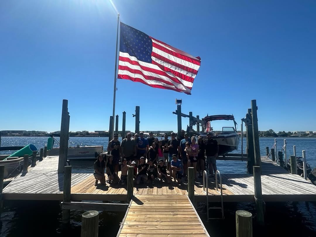 A group gathered on a dock beneath a large American flag on a clear day