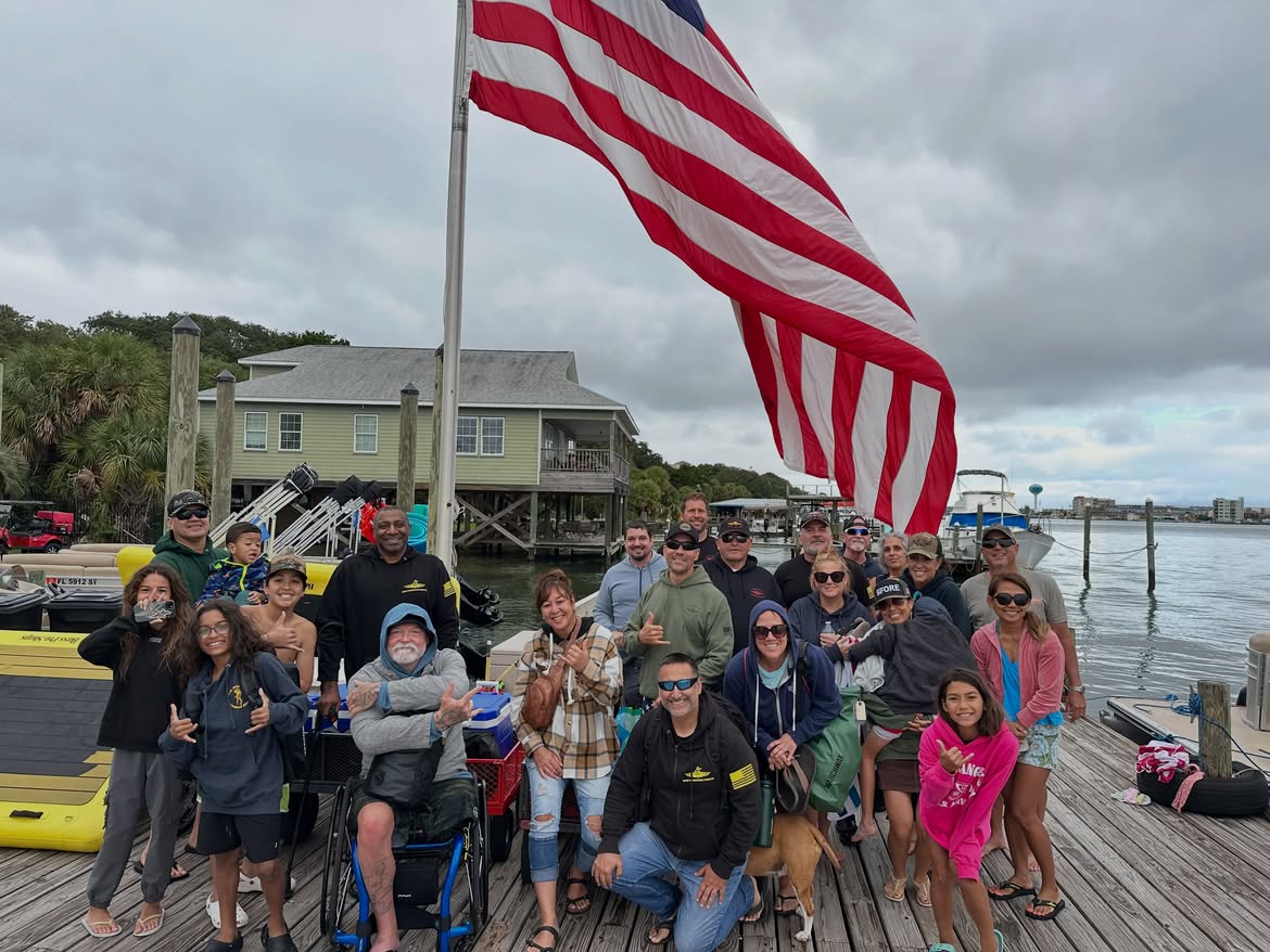 A group of adults and children smiling together on a dock beneath an American flag